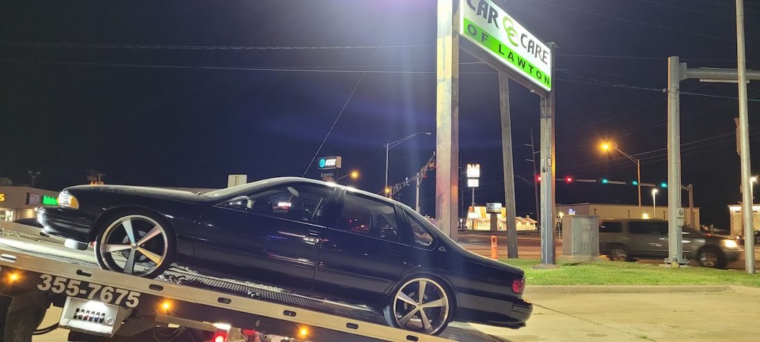 A car loaded on the Car Care of Lawton tow truck at night, beneath the lit shop sign on Cache Road.